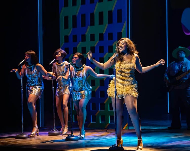 Performer in a gold sequined dress sings centre stage with backing dancers in blue costumes under coloured stage lighting.