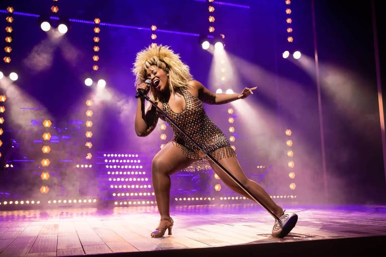 Performer in sequined bodysuit striking a dynamic pose on stage beneath purple and white spotlights.