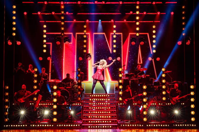 Performer in pink dress centre stage beneath towering neon signs and elaborate red and gold light display.