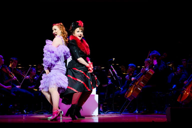 Two performers in lavender and red period costumes dance on stage with an orchestra in the background.