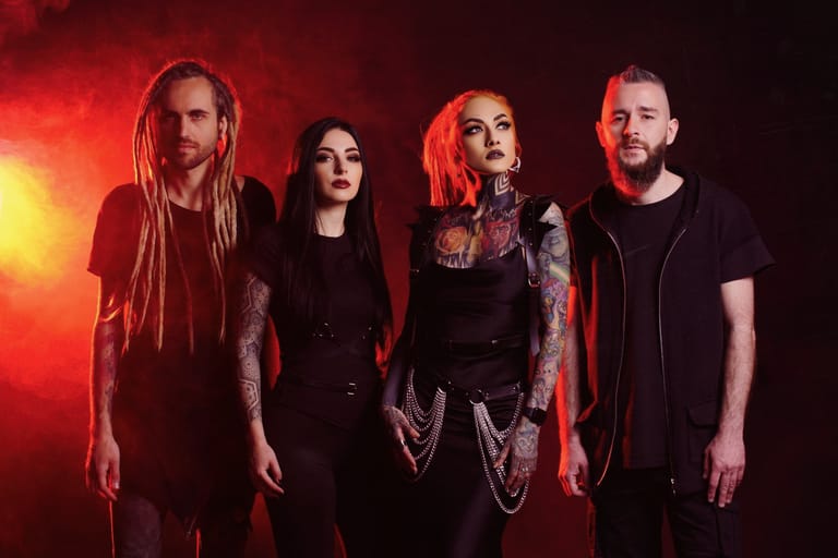 Four band members stand in profile against warm red stage lighting, dressed in dark clothing.