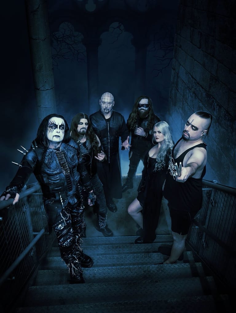 Six musicians in gothic stage makeup and dark clothing stand on a concrete staircase in moody blue lighting.