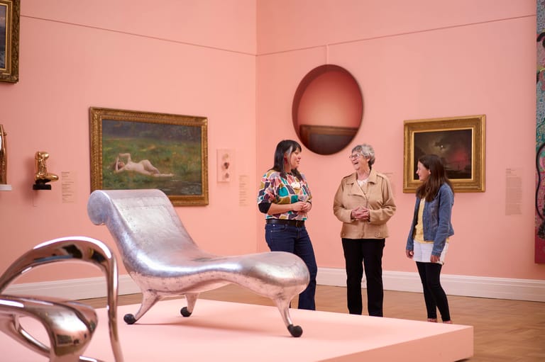 Three people converse in a gallery with pink walls, framed paintings, and a sculptural silver lounge chair in the foreground.