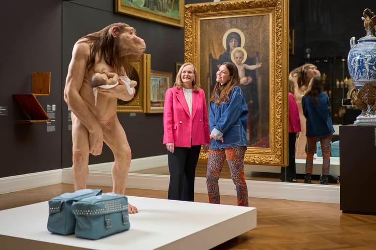 Wooden sculpture of a crouching figure on gallery plinth with visitors viewing framed religious artwork behind.