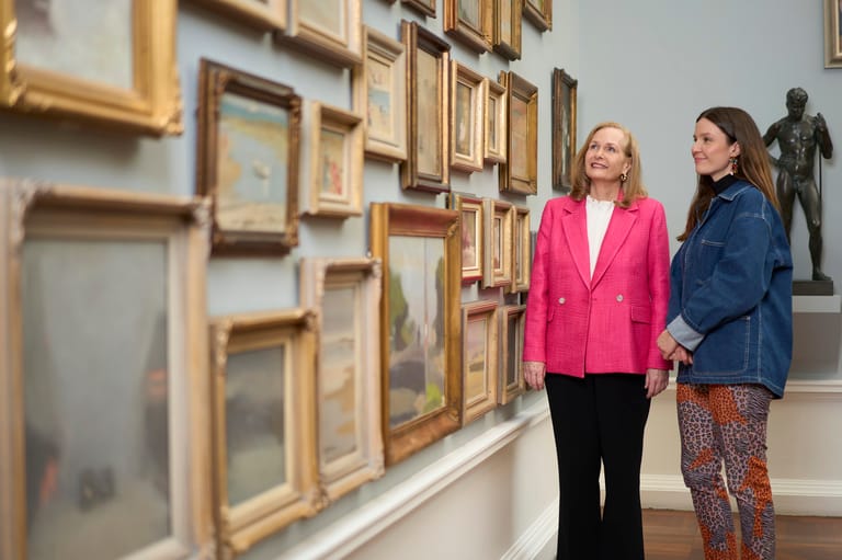 Two women view a gallery wall displaying framed artworks in gold and natural wooden frames.