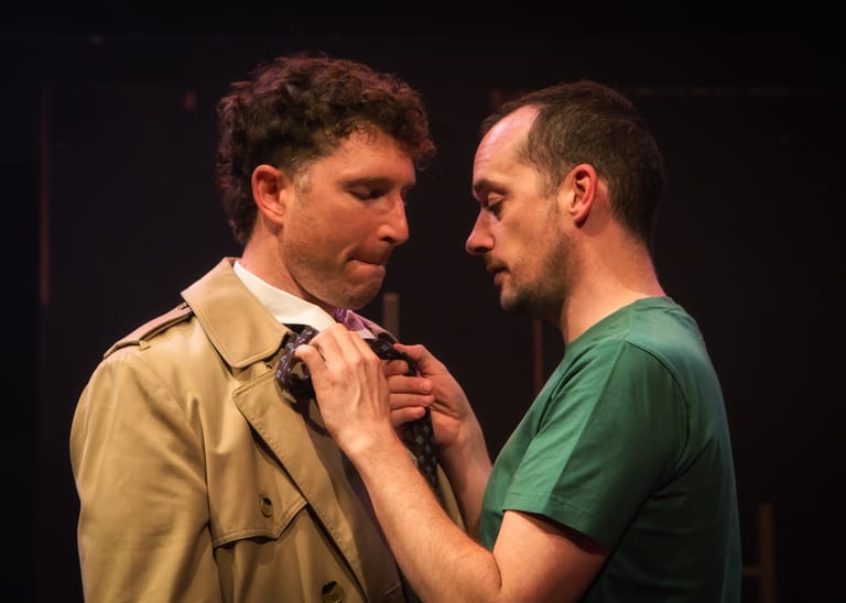 Two men face each other on stage in an intimate dramatic scene under warm lighting.