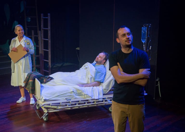 Man in black shirt stands centre stage beside a hospital bed with a patient, whilst a woman in a yellow cardigan watches from stage left.