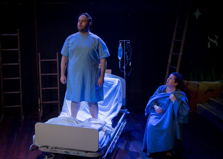 Actor in blue surgical gown stands on hospital bed whilst another performer watches during stage production.