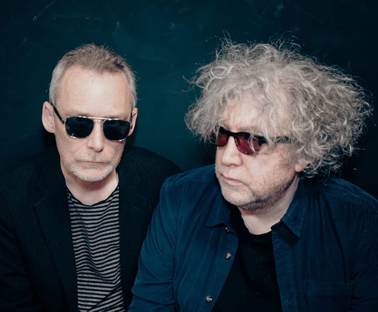 Two musicians wearing sunglasses against a dark teal background.