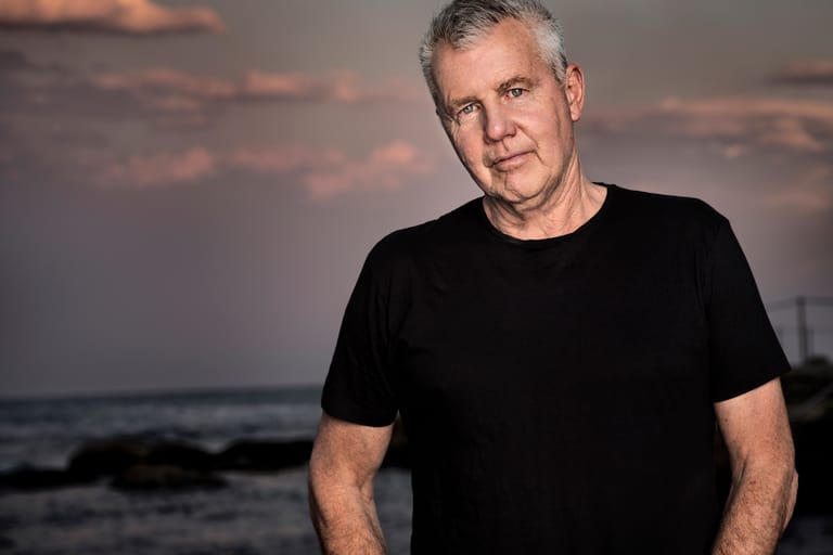 Man in black t-shirt standing against a dusky coastal backdrop with waves visible behind him.