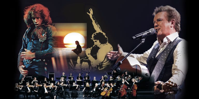 Two musicians perform with a large orchestra under warm stage lighting, featuring a guitarist on the left and a vocalist on the right.