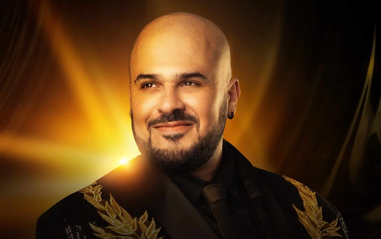 Bald man with beard wearing black and gold jacket, lit by warm golden stage lighting.