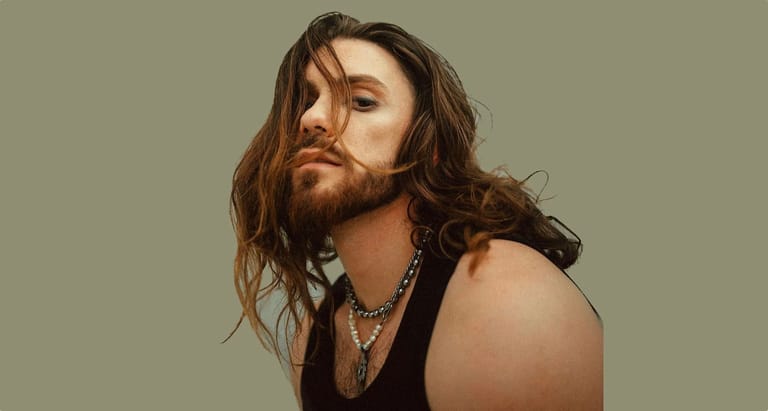 Man with long dark hair and beard wearing a black sleeveless top against a neutral grey background.