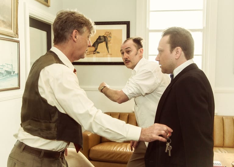 Three men in formal attire examine a golden object during an indoor scene.