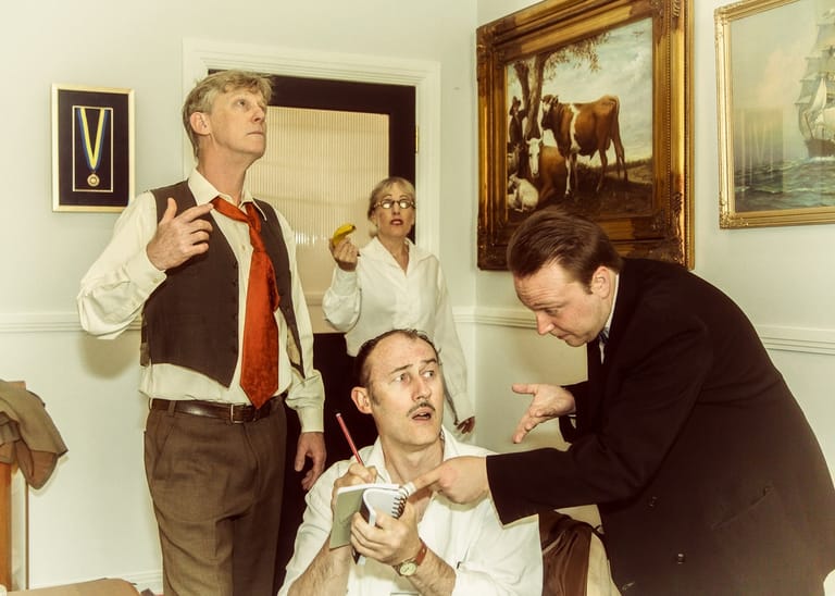 Four men in period clothing perform a comedic scene in a domestic interior setting with framed artwork on the walls.