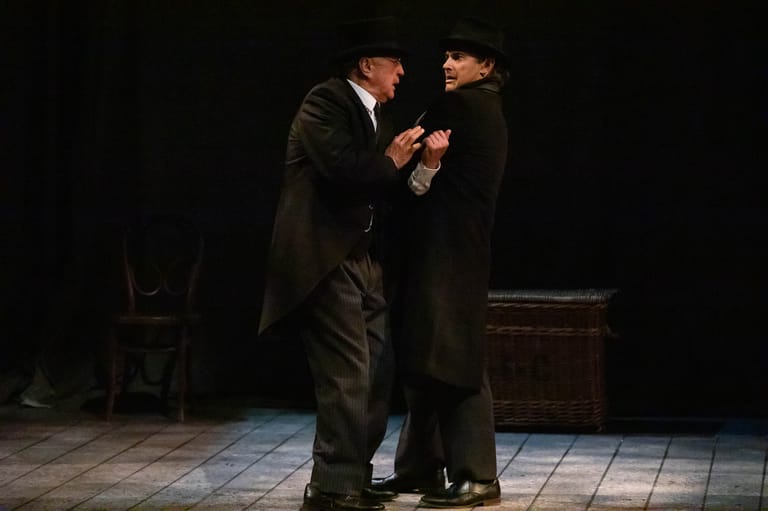 Two men in dark suits engage in conversation on a dimly lit stage set with period furniture.