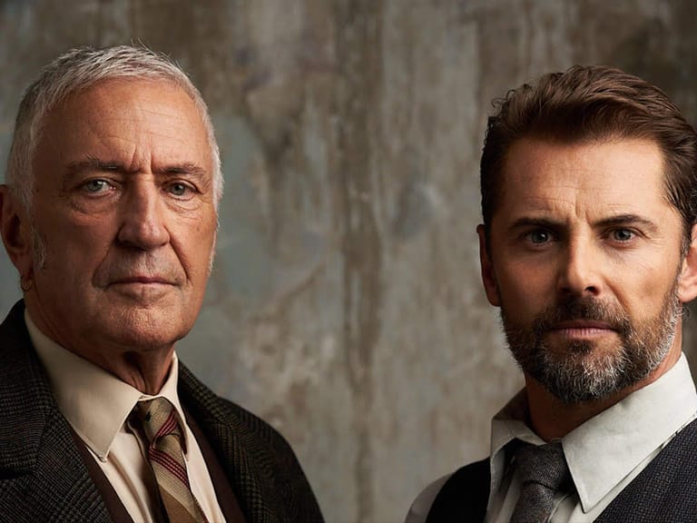 Two men in formal suits and ties stand against a textured grey wall in a studio portrait setting.