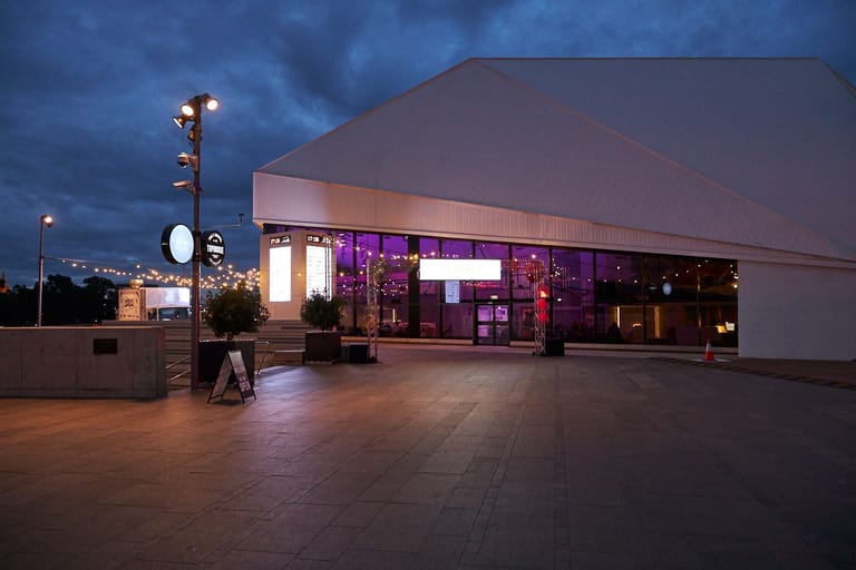 Modern arts venue with illuminated purple and pink signage glows at dusk beneath a dramatic twilight sky.