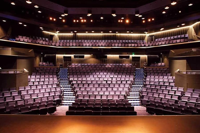 Empty theatre auditorium with purple velvet seats, warm stage lighting, and tiered seating arranged in a semicircle.