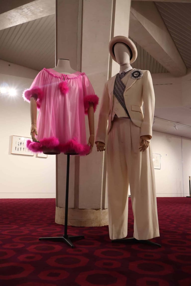 Two mannequins display vintage fashion: a bright pink tulle dress with magenta fur trim and a cream wool coat with brown diagonal stripe detail.