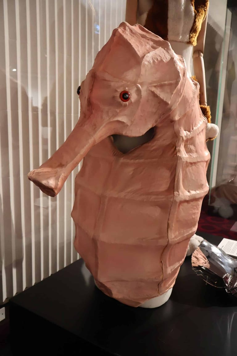 Pink fabric puppet head with elongated snout and circular eyes displayed on a black stand.