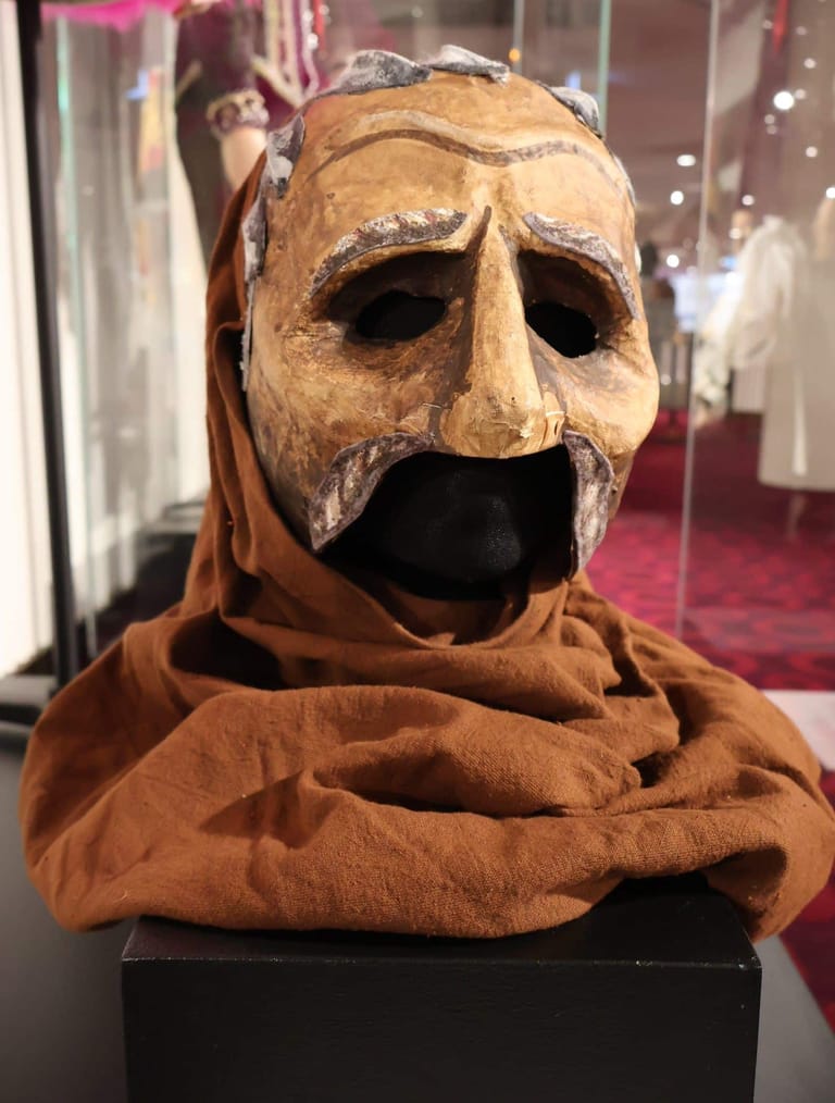 Carved wooden mask with exaggerated features displayed in a museum case with burgundy backdrop.