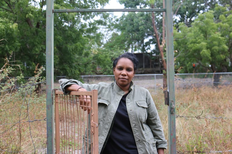 Person in grey jacket standing at open farm gate with dry grass and trees visible behind.