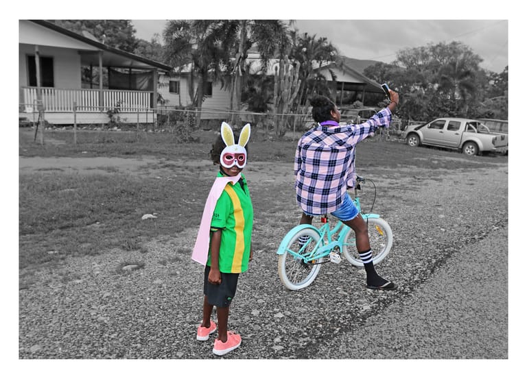 Child in colourful outfit standing next to another child doing a wheelie on a small bike in a rural driveway.