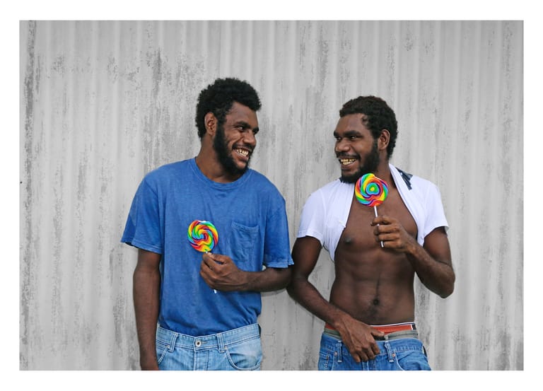 Two men holding lollipops smile at each other against a weathered grey corrugated metal backdrop.