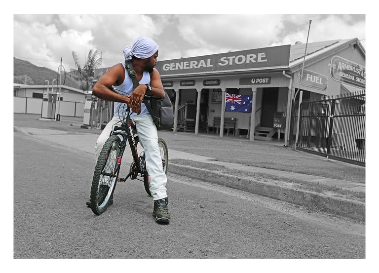 Cyclist in orange shirt and blue cap riding past a heritage general store building.
