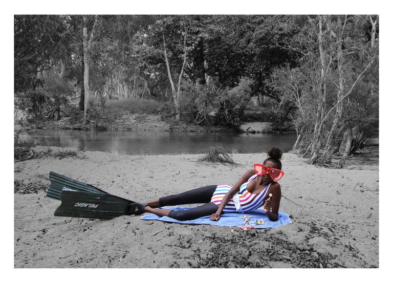 Person in red and blue sitting on sand beside fins, with tree-lined river in background.