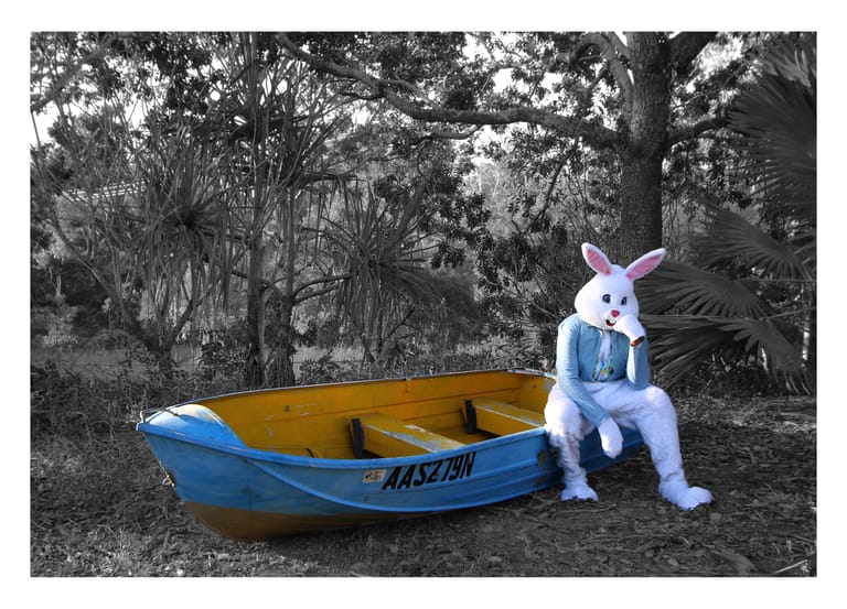 Person in white rabbit costume with blue accents seated beside a yellow and blue rowboat in a wooded setting.