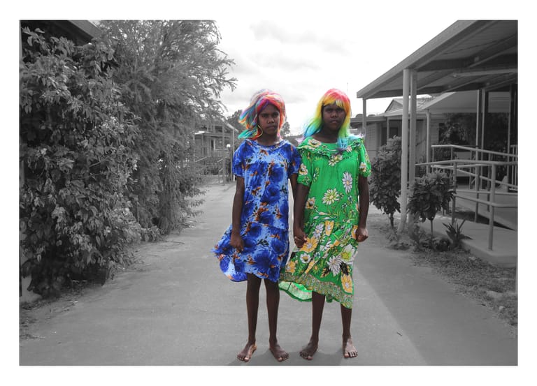 Two people in vibrant blue and green patterned dresses stand together on a residential street lined with trees.