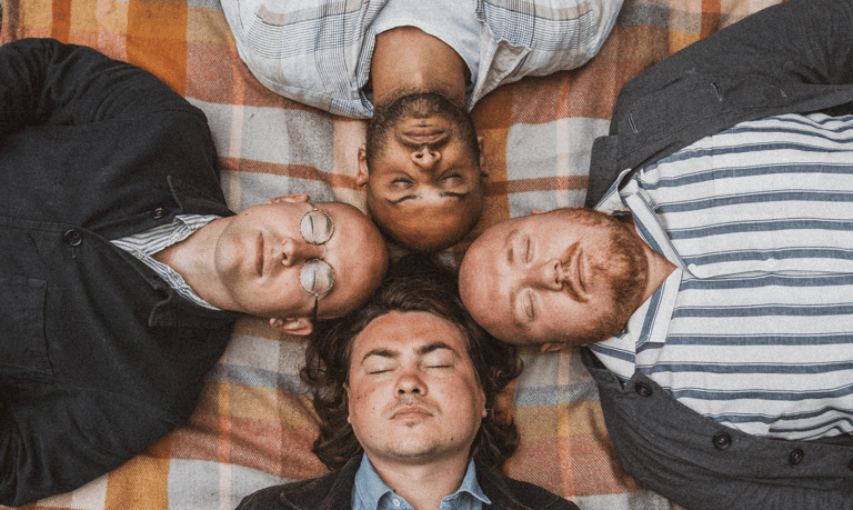 Four people lying on a patterned blanket with heads together, looking down at the camera in a circle formation.