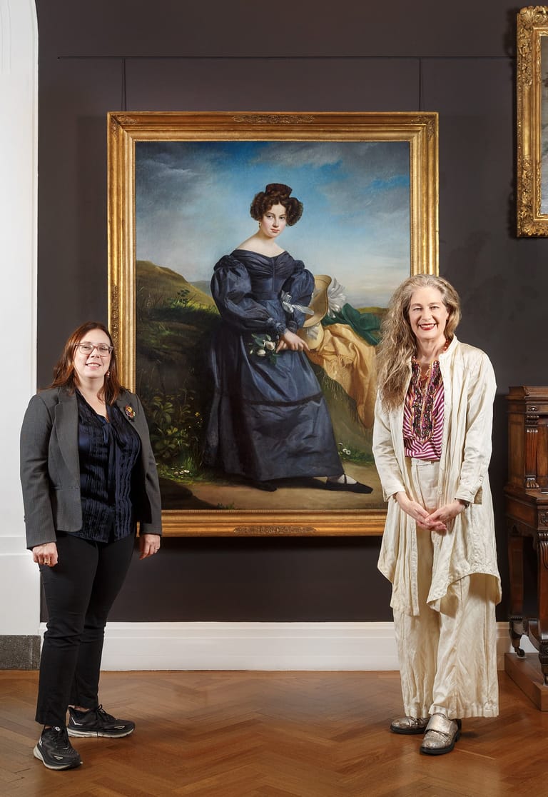 Two women stand beside a large gilt-framed portrait of a woman in a dark blue dress against a landscape background.