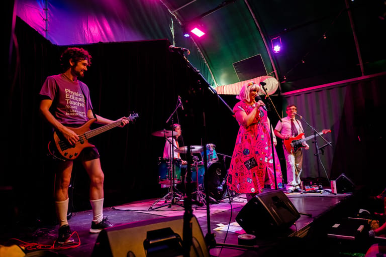 Band performs on stage illuminated by pink and purple stage lighting, with guitarist on left and lead singer centre stage.