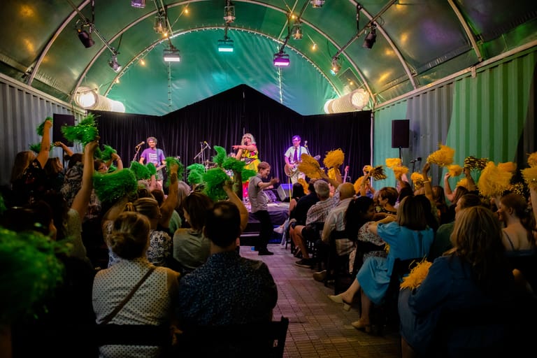 Audience members gather around a stage lit by green and purple lights in an ornate venue during a live performance.