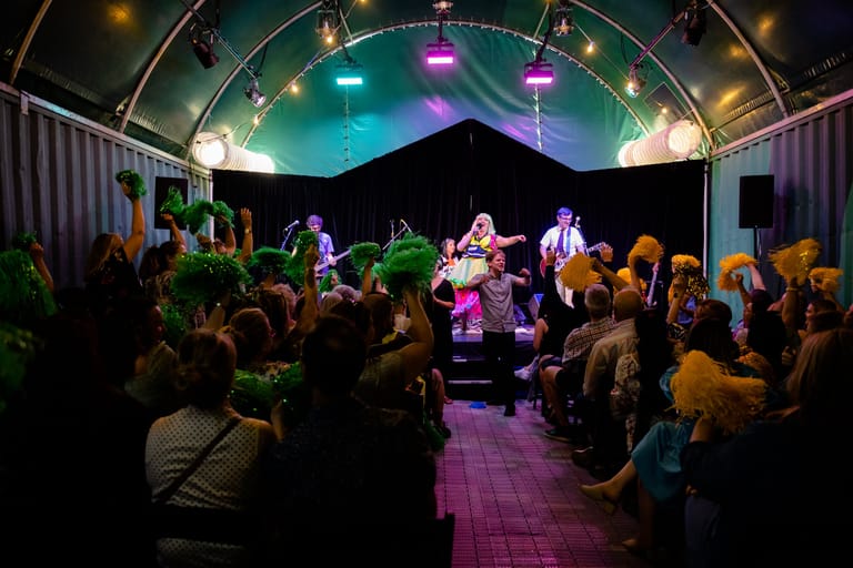 Band performs on an elevated stage beneath coloured lights to a crowded, attentive audience in an intimate indoor venue.