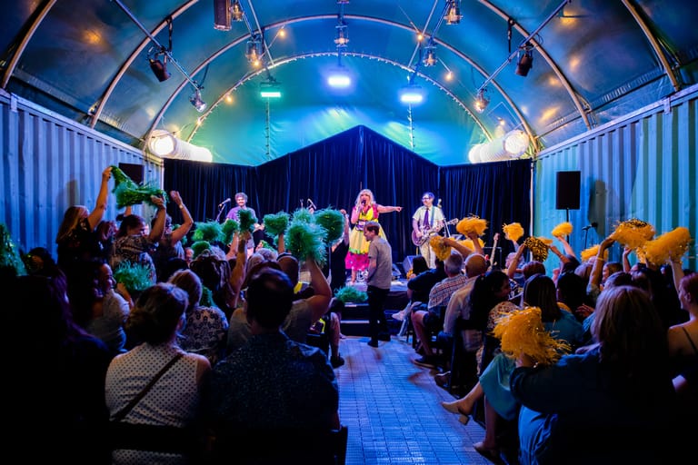 Musicians perform on stage beneath a curved blue-lit venue dome, surrounded by seated audience members.