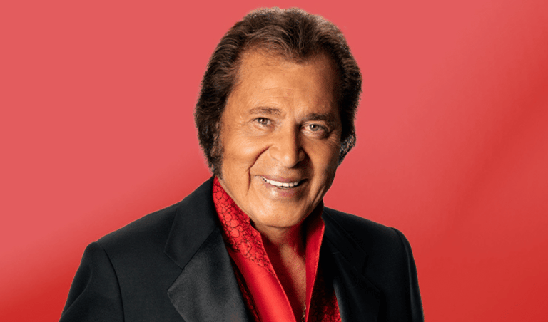 Mature male performer in black jacket and red shirt against red background, smiling at camera.