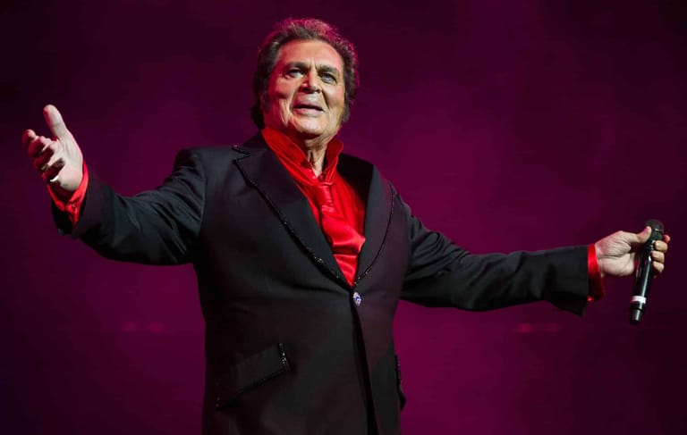 Singer in black jacket and red shirt performing with arms outstretched under purple stage lighting.