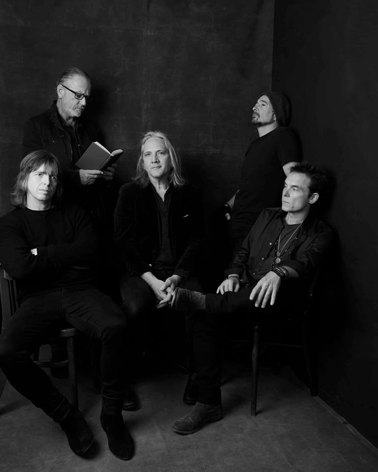 Five musicians posed against a dark backdrop in black and white portrait photography.