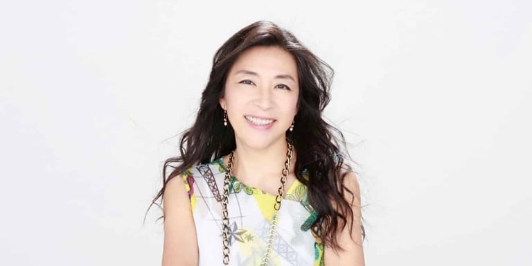 Woman with long dark hair wearing a white sleeveless top and beaded necklace, smiling against a white background.