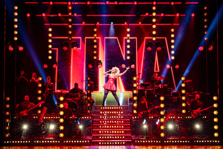 Performer in a sequined outfit stands centre stage beneath glowing neon signs and warm-coloured stage lights during a theatrical production.