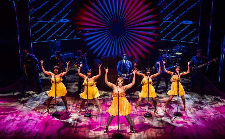 Five performers in yellow dresses dance on a neon-lit stage with geometric backdrop and pink floor lighting.