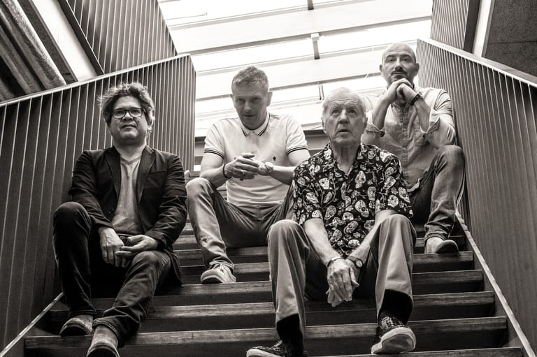 Four musicians sitting on stairs in a black and white photograph, looking toward the camera.