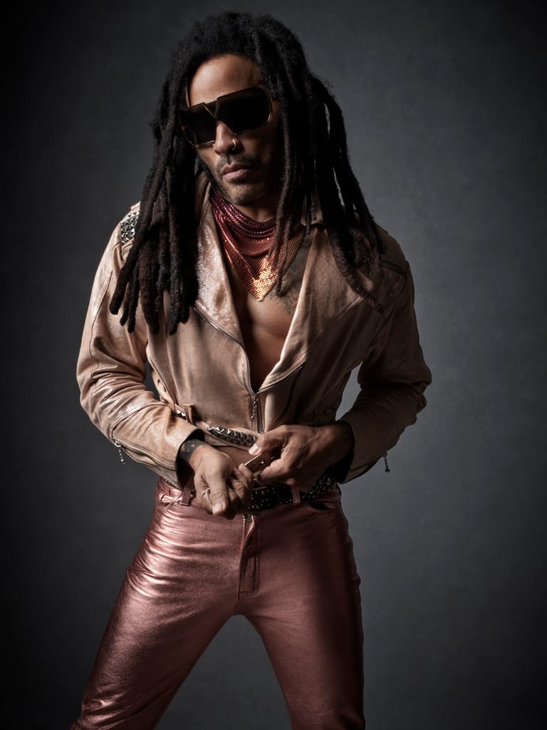 Person with long dreadlocks wearing sunglasses, beige shirt and burgundy pants poses against a dark background.