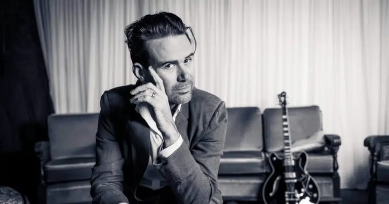 Man in dark shirt speaking on telephone beside electric guitar in studio setting.
