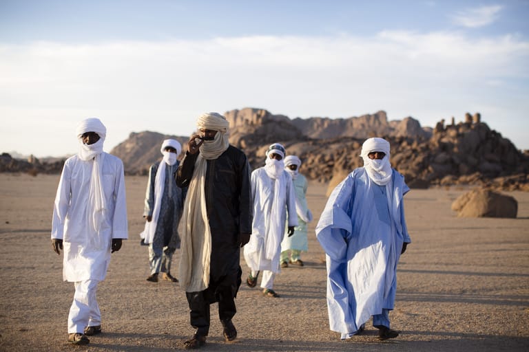 Group of people in traditional robes walking across an arid desert landscape with rocky outcrops behind them.