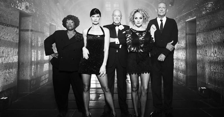Five performers in black formal attire stand posed in a stark, industrial corridor lit by overhead lights.